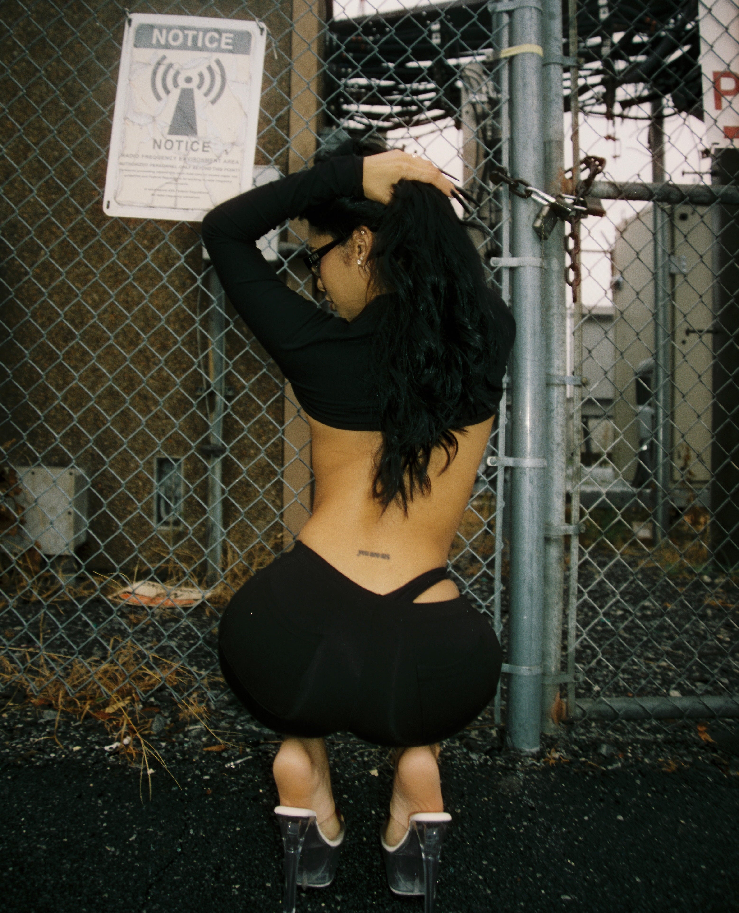 Person wearing a black outfit standing against a chain-link fence with a 'Notice' sign.
